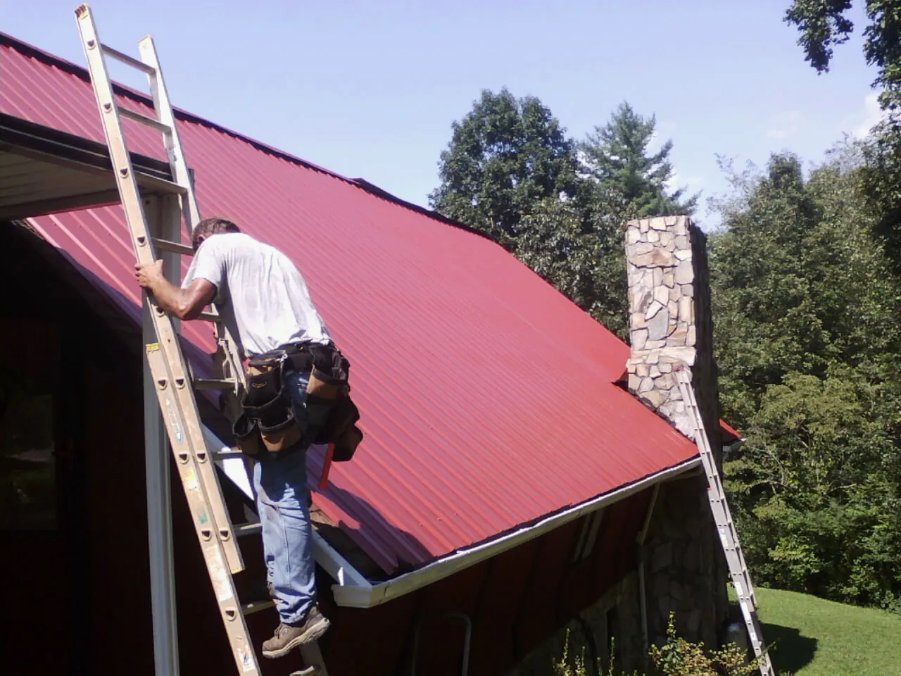 Roofer on a ladder beside a red metal roof and stone chimney — Soffit & Fascia Repair by Roofing Brothers in Harriman
